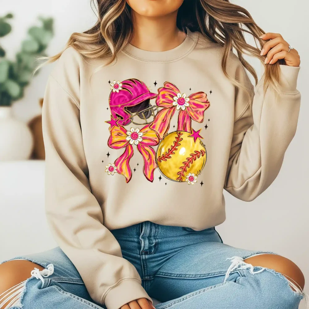 Girl sitting wearing a light brown sweatshirt with a coquette softball design