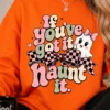 A woman in a bright orange sweatshirt featuring a coquette halloween sublimation typography design that says If you've got it haunt it with a checkered bow and ghost.