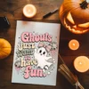 Overhead flat lay of a printed art page featuring the retro pink Halloween design Ghouls just want to have fun with a ghost and disco balls, styled on a wooden tabletop with lit candles and a lit jack-o'-lantern.