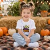 A smiling young girl with hair in pigtails sits cross-legged on a plaid blanket surrounded by pumpkins, wearing a white kids Halloween t-shirt featuring a cute green Frankenstein Monster Munch sublimation design.