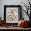 Black ornate picture frame displaying cute Halloween art print on a rustic wood table, flanked by carved jack-o'-lanterns and dark decorative autumn branches.