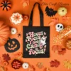 Flat lay of a black canvas tote bag featuring a trendy Hocus Pocus I Need Candy to Focus PNG design, styled on an orange background with spilled candy corn, autumn leaves, a paper bat, and mini pumpkins.