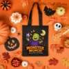 A festive flat lay view of a black Halloween trick-or-treat candy bag featuring a commercial use cute monster sublimation design, surrounded by candy corn, miniature pumpkins, and fall leaves on an orange background.