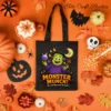 A festive flat lay view of a black Halloween trick-or-treat candy bag featuring a commercial use cute monster sublimation design, surrounded by candy corn, miniature pumpkins, and fall leaves on an orange background.