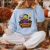 A woman sits cross-legged outdoors on a woven rug holding a mug, wearing comfortable white sweatpants and a light blue oversized crewneck sweatshirt featuring a colorful Halloween candy PNG sublimation design.