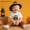 A laughing toddler boy wearing a black witch hat and striped pants sits on a wooden floor next to a lantern, wearing a white infant Halloween shirt with a retro Frankenstein candy digital design.