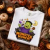 Cozy autumn flat lay mockup of a folded white Comfort Colors sweatshirt featuring a vibrant Halloween DTF graphic of a friendly monster, candy corn, and cute black cats on a dark wooden background with leaves and candles.