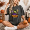 A blonde woman wearing a wide-brimmed hat sits on a boho rug holding a jack-o'-lantern mug, modeling an oversized charcoal gray t-shirt printed with a spooky season digital Frankenstein sublimation PNG.