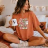 Young woman sitting on a bed in a terracotta t-shirt printed with spooky season PNG art, holding a ghost-shaped coffee mug next to fall bedding.