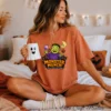 A woman sits cross-legged on a bed holding a white ghost coffee mug, wearing a terracotta orange oversized tee featuring a commercial use cute monster Halloween sublimation design.