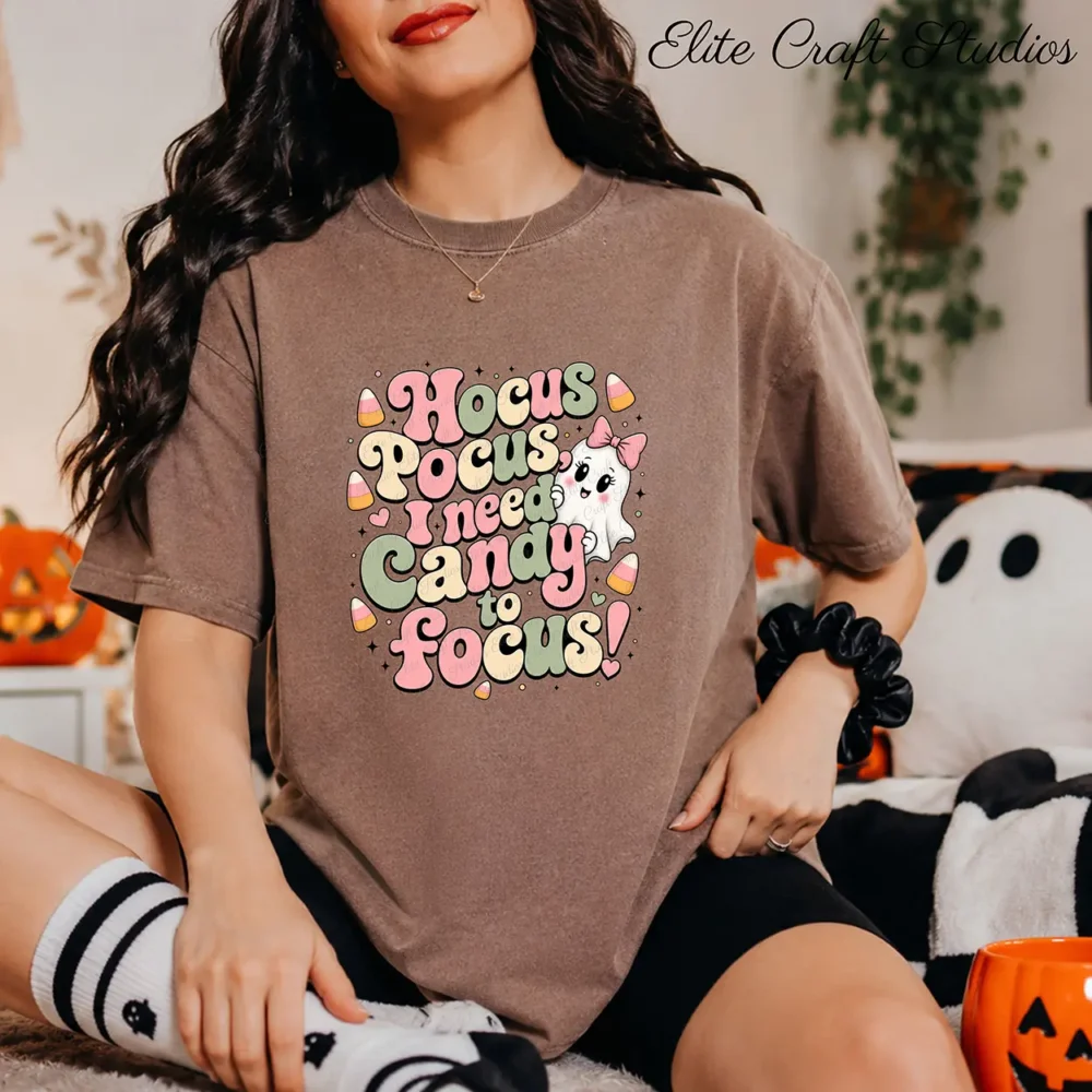 Woman wearing a brown cotton t-shirt with cute Halloween art sublimation print, sitting on the floor with plush ghosts and a pumpkin candy bucket.
