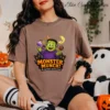 A woman sits on the floor wearing spooky ghost socks and an oversized chocolate brown t-shirt featuring a vibrant Frankenstein Monster Munch sublimation design, next to a festive pumpkin pail.