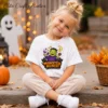 A smiling young girl with a blonde topknot sits cross-legged on outdoor concrete steps, wearing a white kids Halloween t-shirt printed with a cute trick or treat monster PNG graphic.