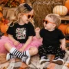 Two young kids sitting together wearing orange heart-shaped sunglasses, showcasing a black t-shirt with trendy Hocus Pocus I Need Candy to Focus PNG design graphic next to a hay bale.
