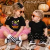 Two young siblings wearing matching heart-shaped sunglasses laugh together on a plaid blanket; the girl wears a black kids Halloween shirt with a retro Frankenstein candy digital design and a pink tutu.