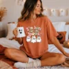 A woman sits casually on a white bed, wearing an oversized rust-colored t-shirt with the retro Halloween PNG design. She is holding a ceramic ghost mug. The background includes string lights and a jack-o'-lantern pillow.