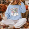 A woman in a light blue oversized sweatshirt with the "I'm Here for the Boos" ghost bow sublimation design sits outdoors on a blanket holding a white mug, surrounded by autumn leaves and hay bales.