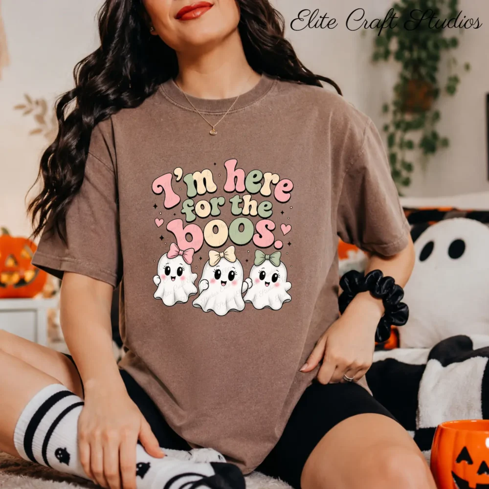 A woman wearing a brown t-shirt and a black scrunchie on her wrist sits on a plush white blanket. The t-shirt has the coquette ghost bow design. A jack-o'-lantern basket is nearby.