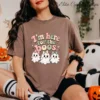 A woman wearing a brown t-shirt and a black scrunchie on her wrist sits on a plush white blanket. The t-shirt has the coquette ghost bow design. A jack-o'-lantern basket is nearby.