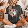 Woman wearing a dark charcoal Halloween t-shirt featuring a cute pink coquette ghost graphic while holding a pumpkin mug.