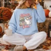 Model sitting outside among autumn leaves wearing a light blue crewneck sweatshirt with the retro pink ghost graphic.