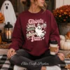 Woman sitting outdoors in a maroon Halloween sweatshirt printed with a pink coquette ghost design and coffee cup.