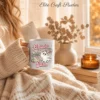 Cozy sweater-clad hands holding a white coffee mug printed with the cute pink coquette Halloween ghost graphic.