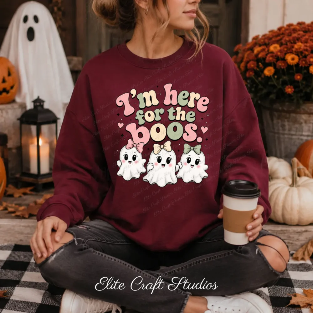 A woman sits cross-legged on a porch wearing a maroon sweatshirt printed with the "I'm Here for the Boos" retro ghost PNG design, holding a coffee cup next to autumn flowers and white pumpkins.