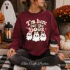A woman sits cross-legged on a porch wearing a maroon sweatshirt printed with the "I'm Here for the Boos" retro ghost PNG design, holding a coffee cup next to autumn flowers and white pumpkins.
