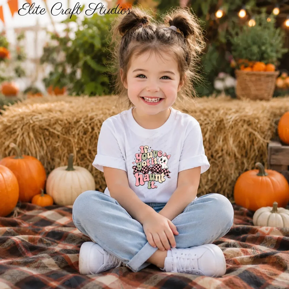 A smiling young girl sitting cross-legged on a plaid blanket at a pumpkin patch wearing a white graphic t-shirt
