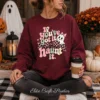 A woman sitting cross-legged on a porch holding a takeout coffee cup while wearing a maroon crewneck sweatshirt