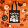 Flat lay view of a black tote bag featuring the spooky cute "I'm Here for the Boos" pastel Halloween PNG, surrounded by fall leaves, candy corn, a mini bat, and mini decorative pumpkins on an orange background.
