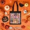 Black canvas tote bag printed with the Creepy and Cute Halloween graphic laid flat on an orange background with candy corn and fall decor.