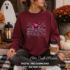 Woman seated outdoors wearing a maroon crewneck sweatshirt with the pink spooky cowgirl print, holding a coffee cup beside fall mums and pumpkins.