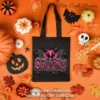 Black canvas tote bag printed with the pink spooky graphic, displayed on a vibrant orange surface with candy corn and fall leaves.