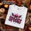 White t-shirt flat lay featuring the pink spooky cowgirl graphic, surrounded by autumn leaves, lit candle, and mini pumpkins.