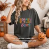 Smiling woman sitting cross-legged wearing a charcoal gray t-shirt featuring the Spooky Season design, holding an orange pumpkin mug.