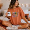 Young woman sitting on a cozy bed holding a ghost mug, wearing a rust orange oversized t-shirt printed with the colorful dripping Spooky Season artwork.