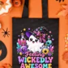 Black reusable tote bag printed with the Feeling Wickedly Awesome ghost design, flat-laid on an orange backdrop surrounded by Halloween decor, candy corn, and a black felt bat.