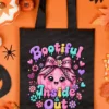 Flatlay top-down view of a black tote bag featuring the Bootiful Inside and Out graphic, surrounded by autumn leaves and Halloween decor.
