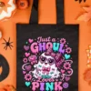 Black canvas tote bag printed with the Just a Ghoul Who Loves Pink design, laid flat on an orange surface with Halloween decor, autumn leaves, and candy corn.