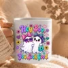 Hand holding a white ceramic mug printed with the colorful Hey Boo ghost design against a soft knit blanket background.