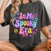 Smiling blonde woman sitting on the floor wearing a charcoal grey oversized t-shirt printed with the In My Spooky Era retro Halloween design.