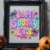 Ornate black picture frame displaying the In My Spooky Era colorful print sitting on a wooden mantel between two carved jack-o'-lantern pumpkins.