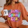 Woman sitting in bed wearing an oversized terracotta orange t-shirt with the In My Spooky Era graphic while holding a white ghost-shaped ceramic mug.