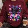 Person sitting cross-legged wearing a maroon pullover sweater printed with resting witch face bat graphic and holding coffee.