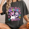 Smiling woman sitting down wearing a dark grey t-shirt featuring the colorful You Look Ghoulishly Good ghost graphic design.
