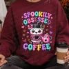 Woman sitting in a cozy dark maroon crewneck sweatshirt with the Spookily Obsessed with Coffee print, holding a disposable coffee cup.