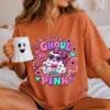 Model seated on a cozy bed wearing a terracotta orange t-shirt printed with the pink ghost design, holding a white ghost mug.