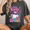 Smiling woman sitting cross-legged wearing a charcoal gray t-shirt printed with the Too Cute to Spook ghost artwork while holding a pumpkin mug.